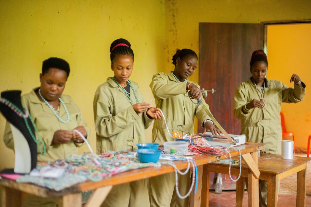 Women in uniforms creating beaded jewelry in an indoor craft workshop.