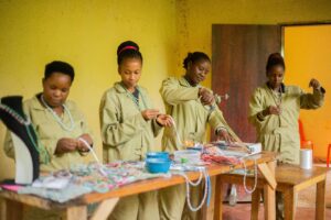 Women in uniforms creating beaded jewelry in an indoor craft workshop.